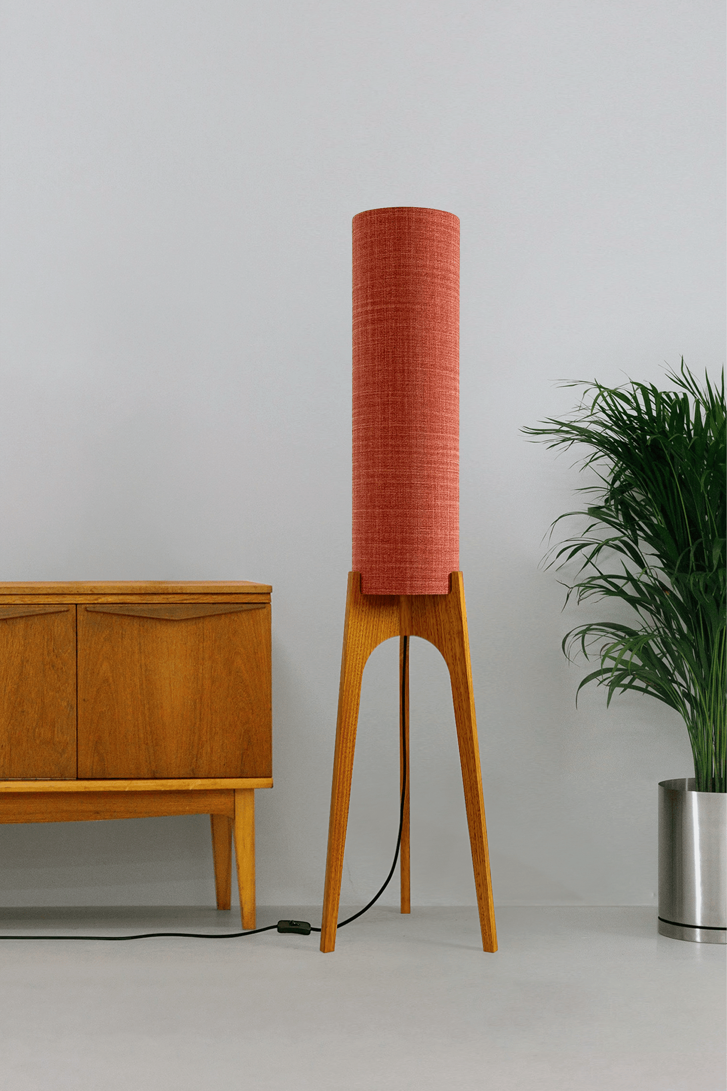 Chili - Tall Archie Rocket floor lamp with red cylindrical shade and wooden base next to a wooden cabinet and plant.