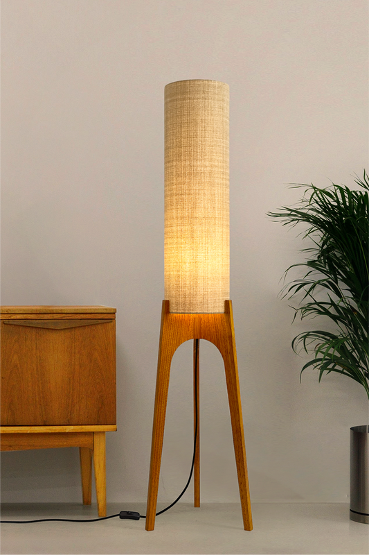 The Linen Archie Rocket Floor Lamp by Retro Duo, featuring a handcrafted wooden base and beige cylindrical shade, styled beside a wooden cabinet and plant against a light grey background.