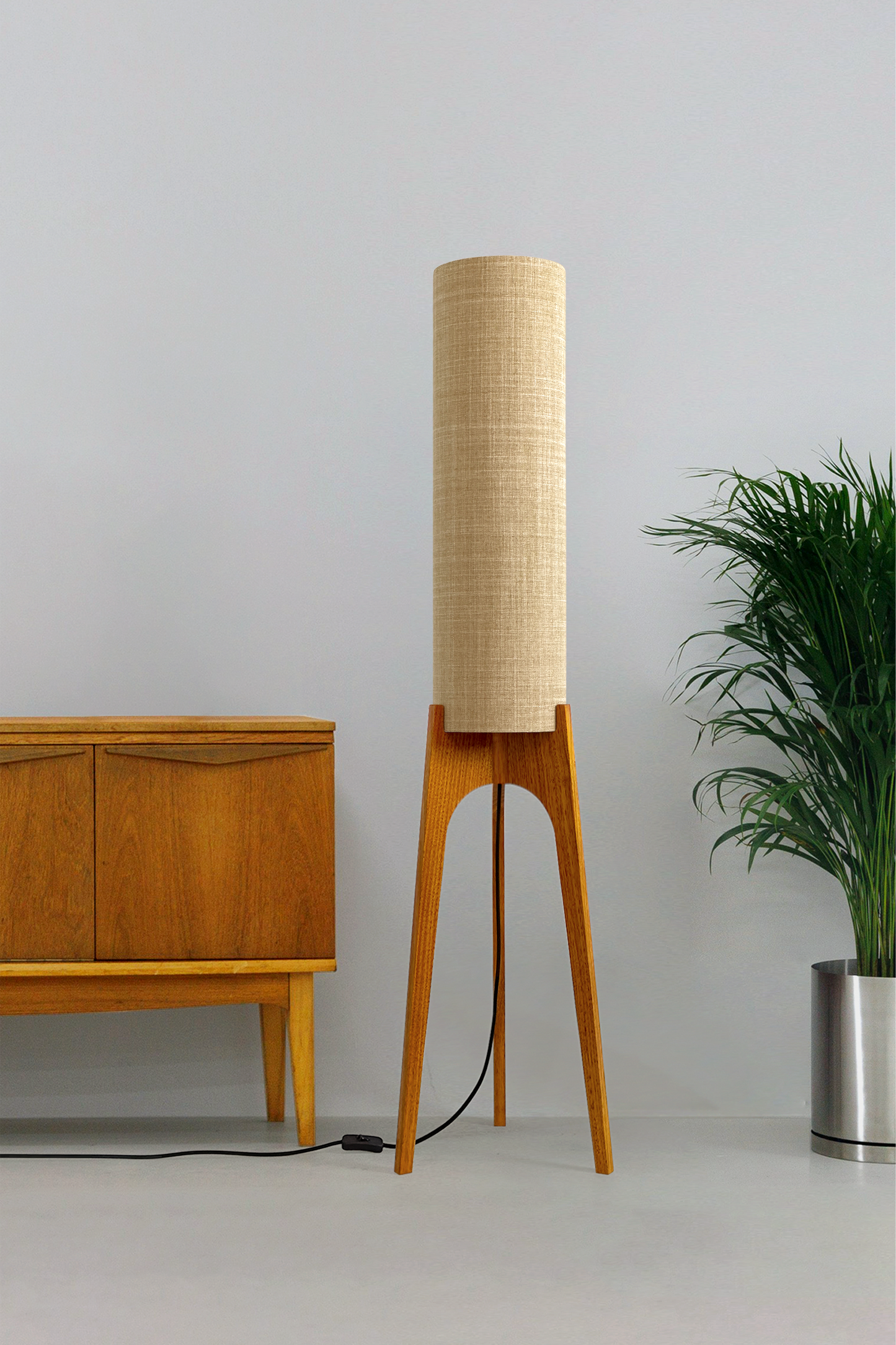 The Linen Archie Rocket Floor Lamp by Retro Duo, featuring a handcrafted wooden base and beige cylindrical shade, styled beside a wooden cabinet and plant against a light grey background.