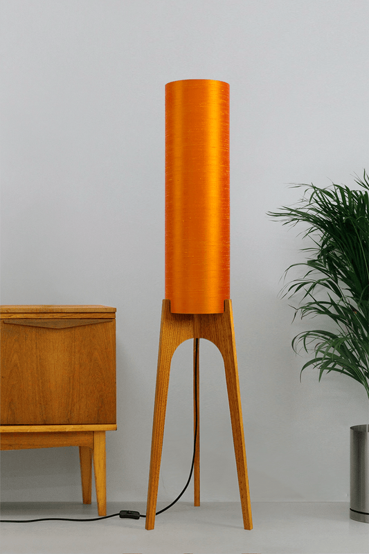 Wooden Rocket Floor Lamp with Orange cylindrical handmade lampshade with premium raw silk, next to a wooden cabinet and plant on a light grey background