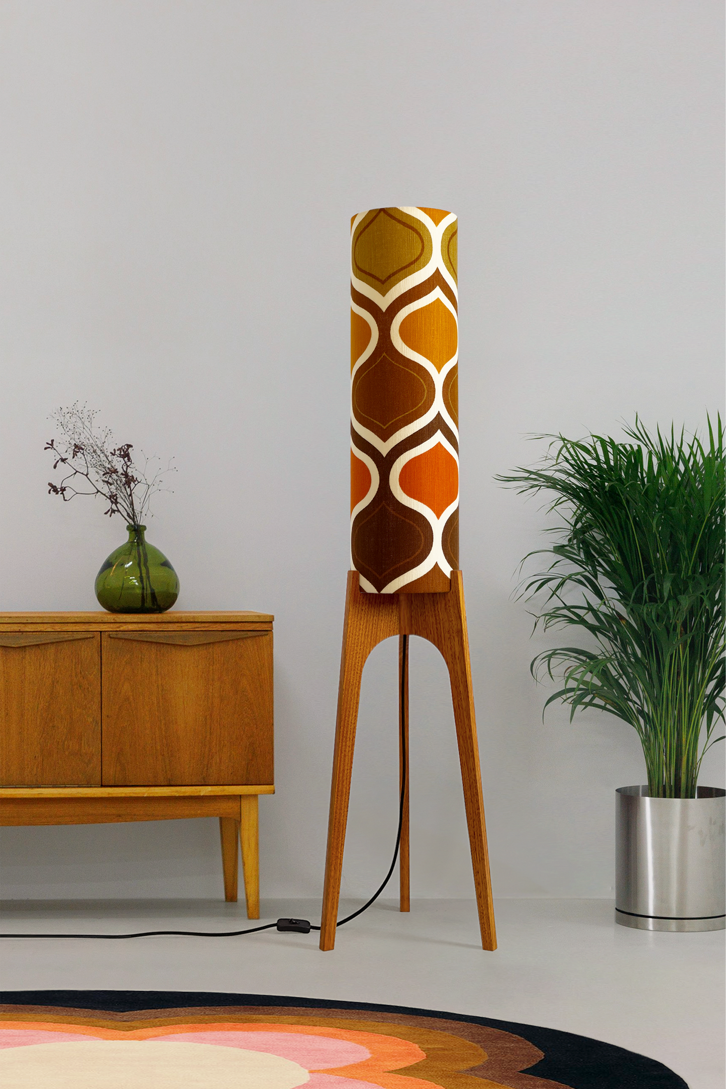 The Tangerine Archie Rocket Floor Lamp is a playful nod to the geometric patterns of the 1970s. Handcrafted from authentic vintage fabric and hardwood oak with a patterned shade in a room setting with a cabinet and plant.