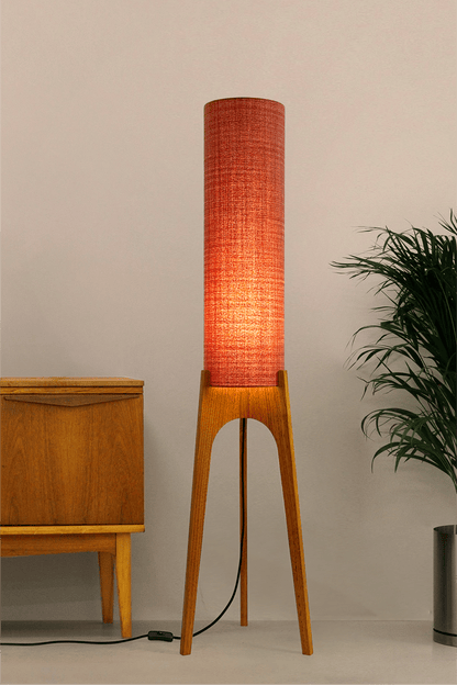 Chili - Tall Archie Rocket floor lamp with red cylindrical shade and wooden base next to a wooden cabinet and plant.