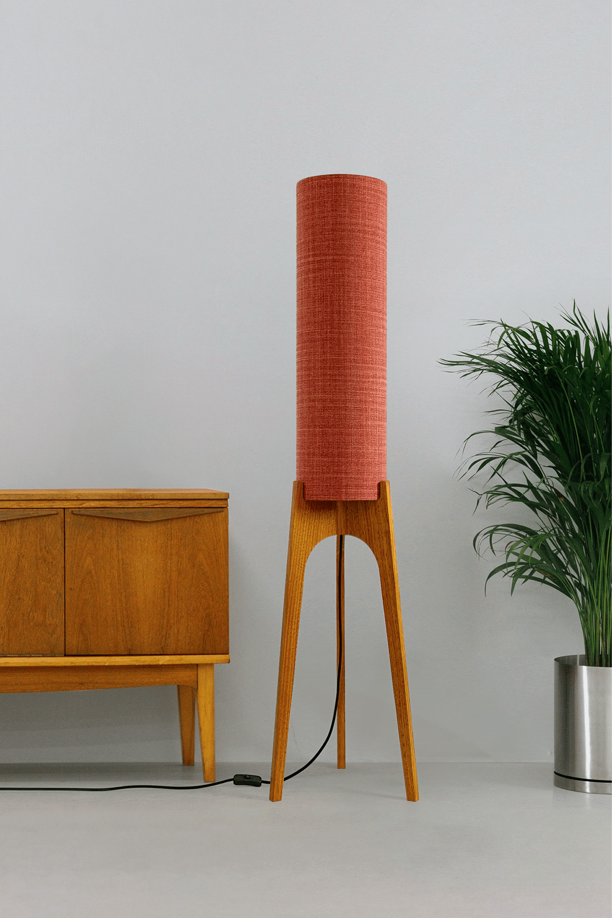 Chili - Tall Archie Rocket floor lamp with red cylindrical shade and wooden base next to a wooden cabinet and plant.