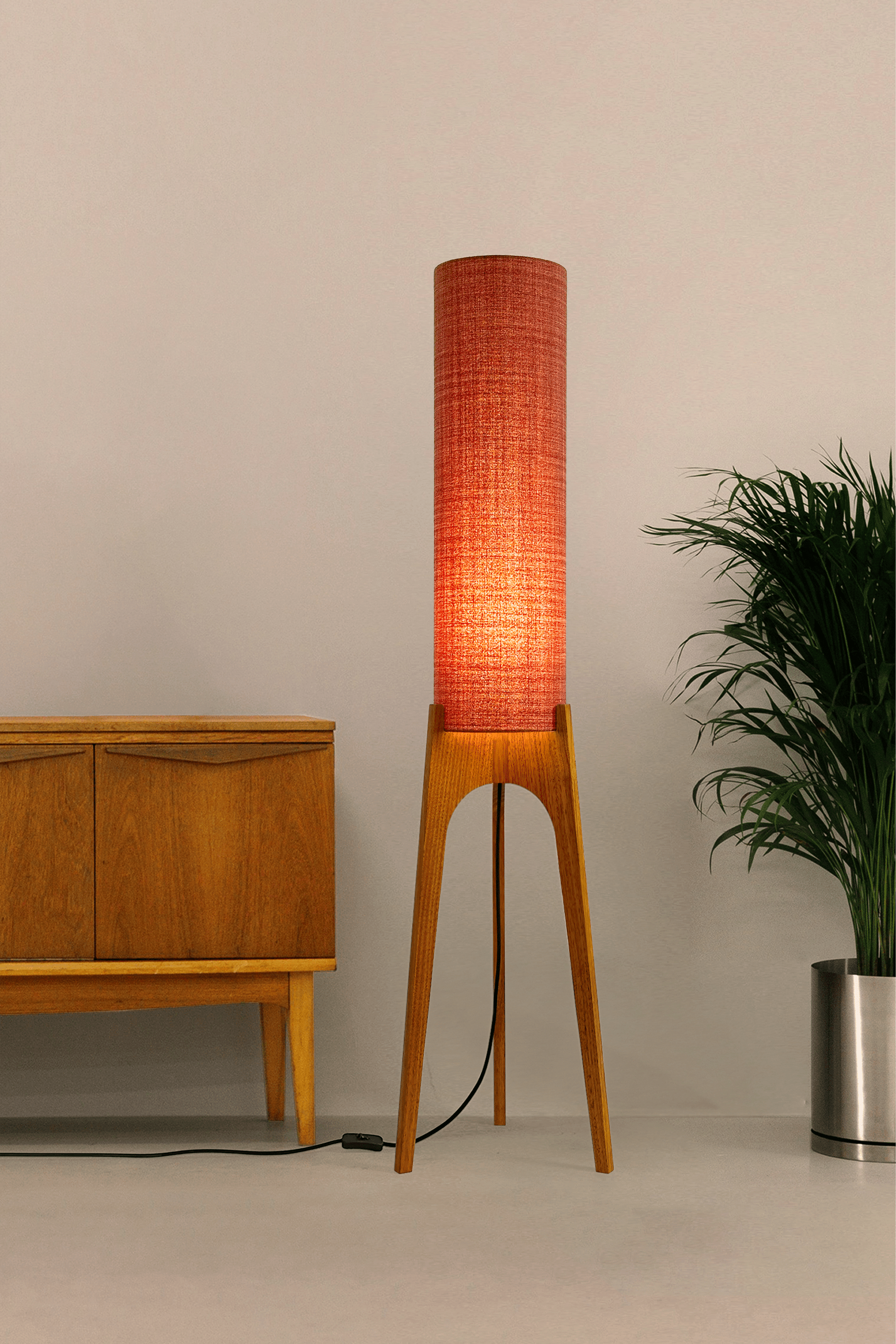 Chili - Tall Archie Rocket floor lamp with red cylindrical shade and wooden base next to a wooden cabinet and plant.