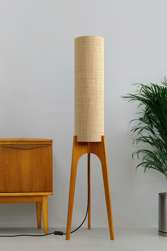 The Linen Archie Rocket Floor Lamp by Retro Duo, featuring a handcrafted wooden base and beige cylindrical shade, styled beside a wooden cabinet and plant against a light grey background.