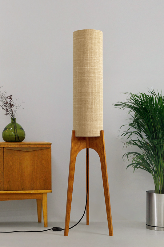 The Linen Archie Rocket Floor Lamp by Retro Duo, featuring a handcrafted wooden base and beige cylindrical shade, styled beside a wooden cabinet and plant against a light grey background.