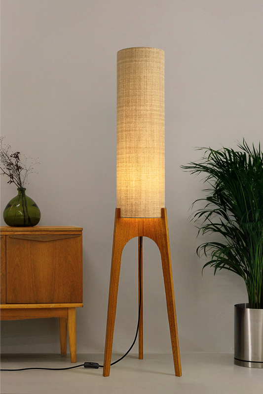 The Linen Archie Rocket Floor Lamp by Retro Duo, featuring a handcrafted wooden base and beige cylindrical shade, styled beside a wooden cabinet and plant against a light grey background.