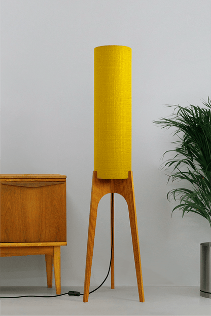 Wooden floor lamp with a yellow handmade lampshade in vintage woven fabric next to a wooden cabinet and a plant on a beige background
