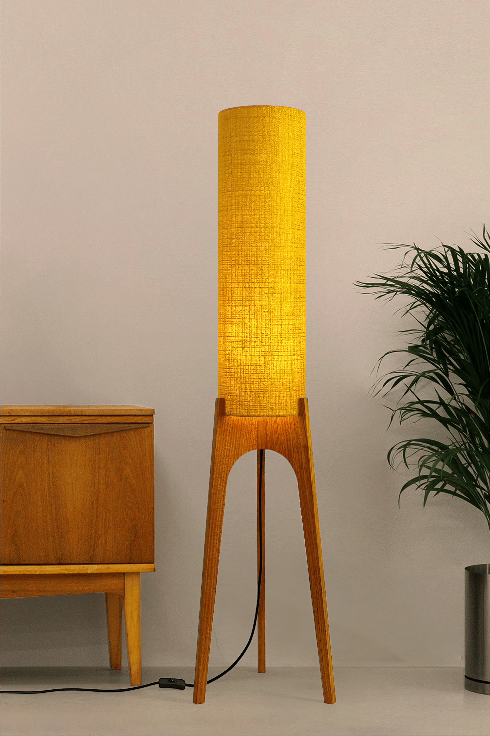 Wooden floor lamp with a yellow handmade lampshade in vintage woven fabric next to a wooden cabinet and a plant on a beige background