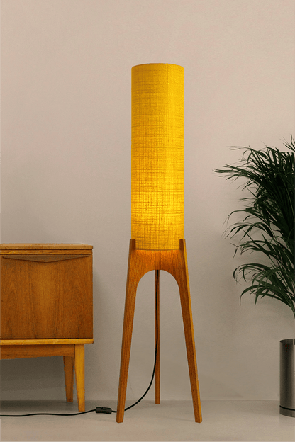 Wooden floor lamp with a yellow handmade lampshade in vintage woven fabric next to a wooden cabinet and a plant on a beige background