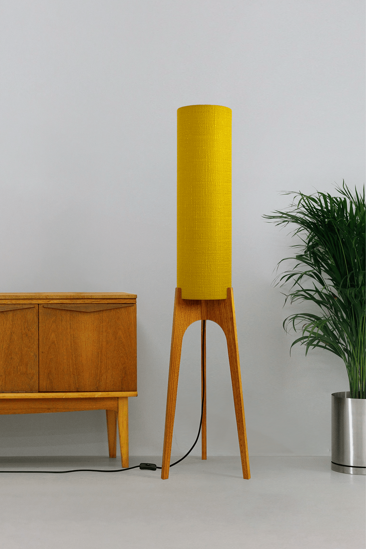 Wooden floor lamp with a yellow handmade lampshade in vintage woven fabric next to a wooden cabinet and a plant on a beige background