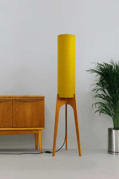 Wooden floor lamp with a yellow handmade lampshade in vintage woven fabric next to a wooden cabinet and a plant on a beige background