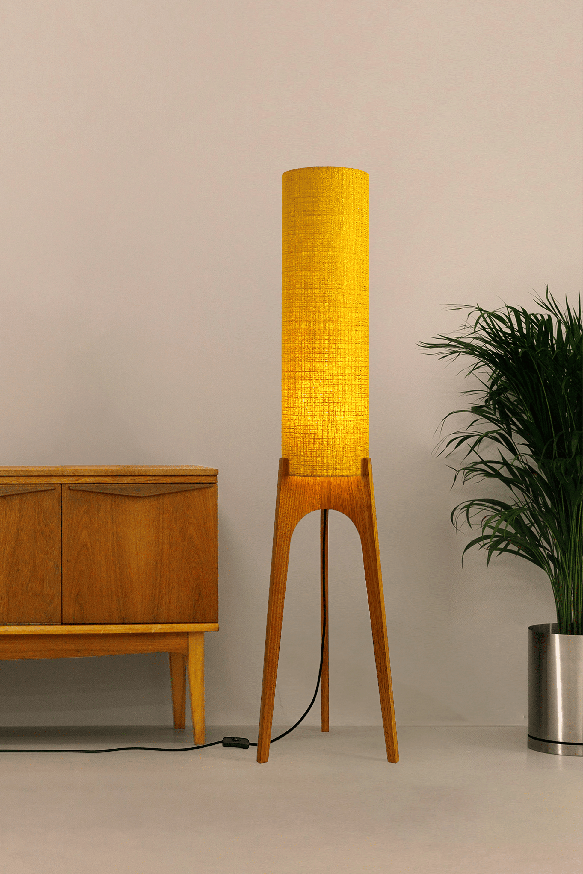 Wooden floor lamp with a yellow handmade lampshade in vintage woven fabric next to a wooden cabinet and a plant on a beige background