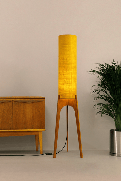 Wooden floor lamp with a yellow handmade lampshade in vintage woven fabric next to a wooden cabinet and a plant on a beige background