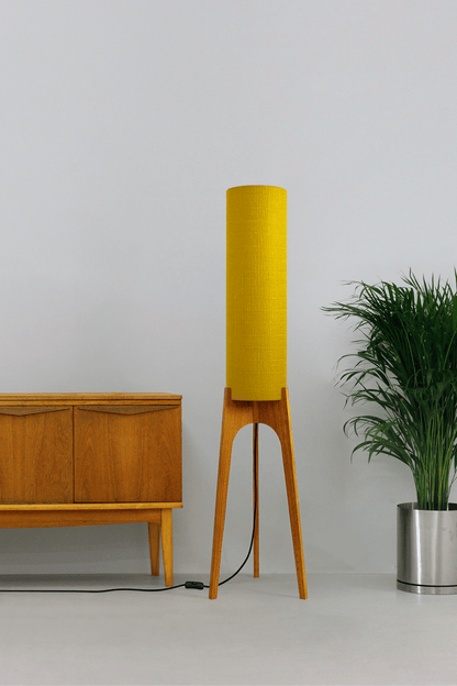 Wooden floor lamp with a yellow handmade lampshade in vintage woven fabric next to a wooden cabinet and a plant on a beige background