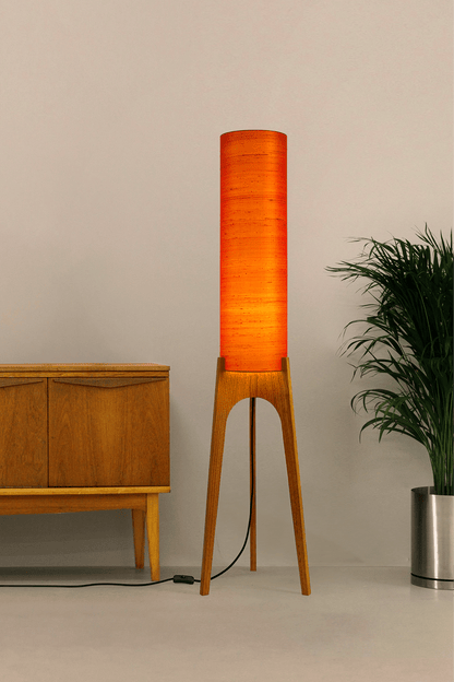 Wooden Rocket Floor Lamp with Orange cylindrical handmade lampshade with premium raw silk, next to a wooden cabinet and plant on a light grey background
