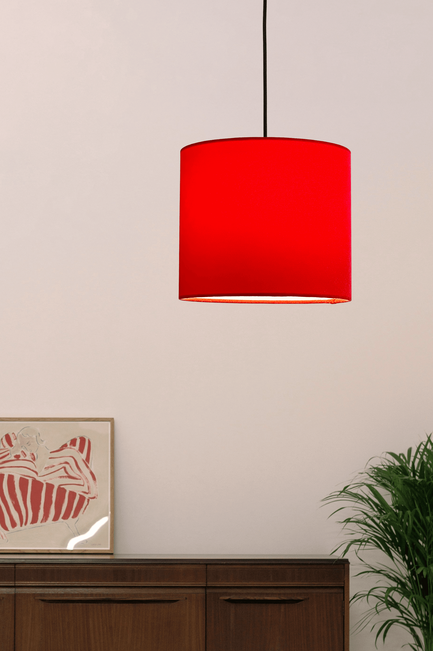 Red pendant lamp hanging against a light wall with a piece of art and plant in the background.