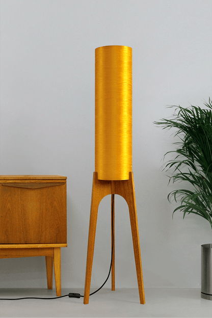 Wooden Rocket Floor Lamp with Saffron Yellow cylindrical handmade lampshade in premium raw silk, next to a wooden cabinet and plant on a light grey background