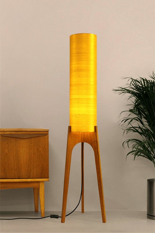 Wooden Rocket Floor Lamp with Saffron Yellow cylindrical handmade lampshade in premium raw silk, next to a wooden cabinet and plant on a light grey background