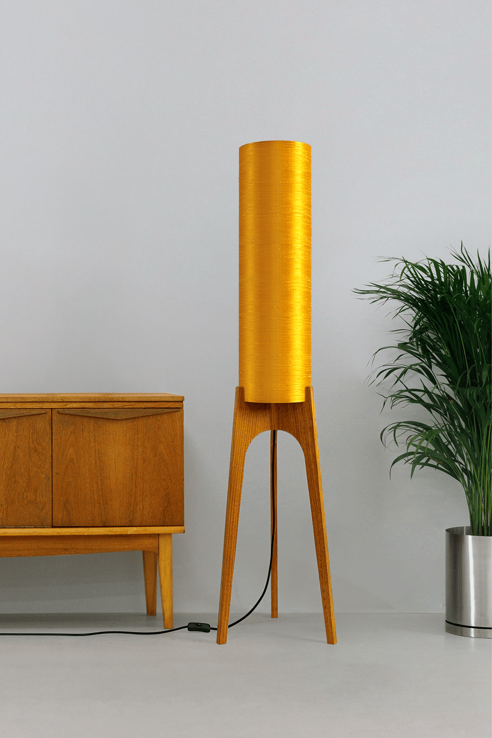 Wooden Rocket Floor Lamp with Saffron Yellow cylindrical handmade lampshade in premium raw silk, next to a wooden cabinet and plant on a light grey background