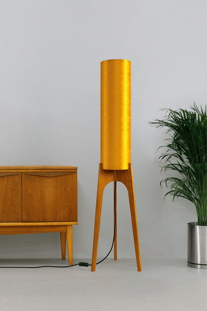 Wooden Rocket Floor Lamp with Saffron Yellow cylindrical handmade lampshade in premium raw silk, next to a wooden cabinet and plant on a light grey background