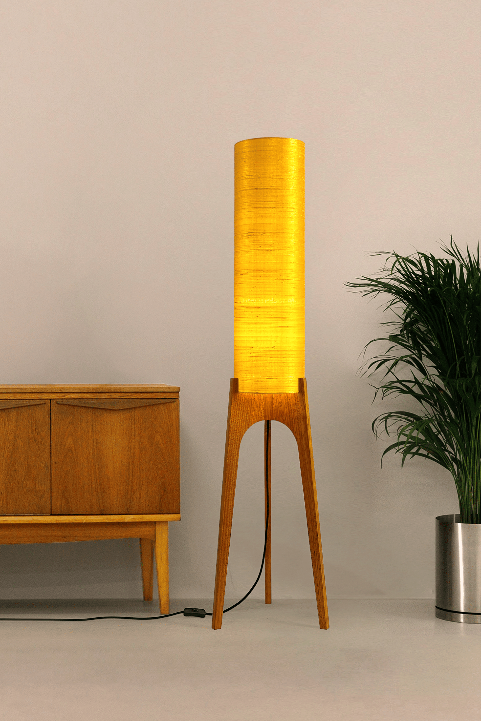 Wooden Rocket Floor Lamp with Saffron Yellow cylindrical handmade lampshade in premium raw silk, next to a wooden cabinet and plant on a light grey background