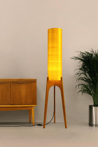 Wooden Rocket Floor Lamp with Saffron Yellow cylindrical handmade lampshade in premium raw silk, next to a wooden cabinet and plant on a light grey background