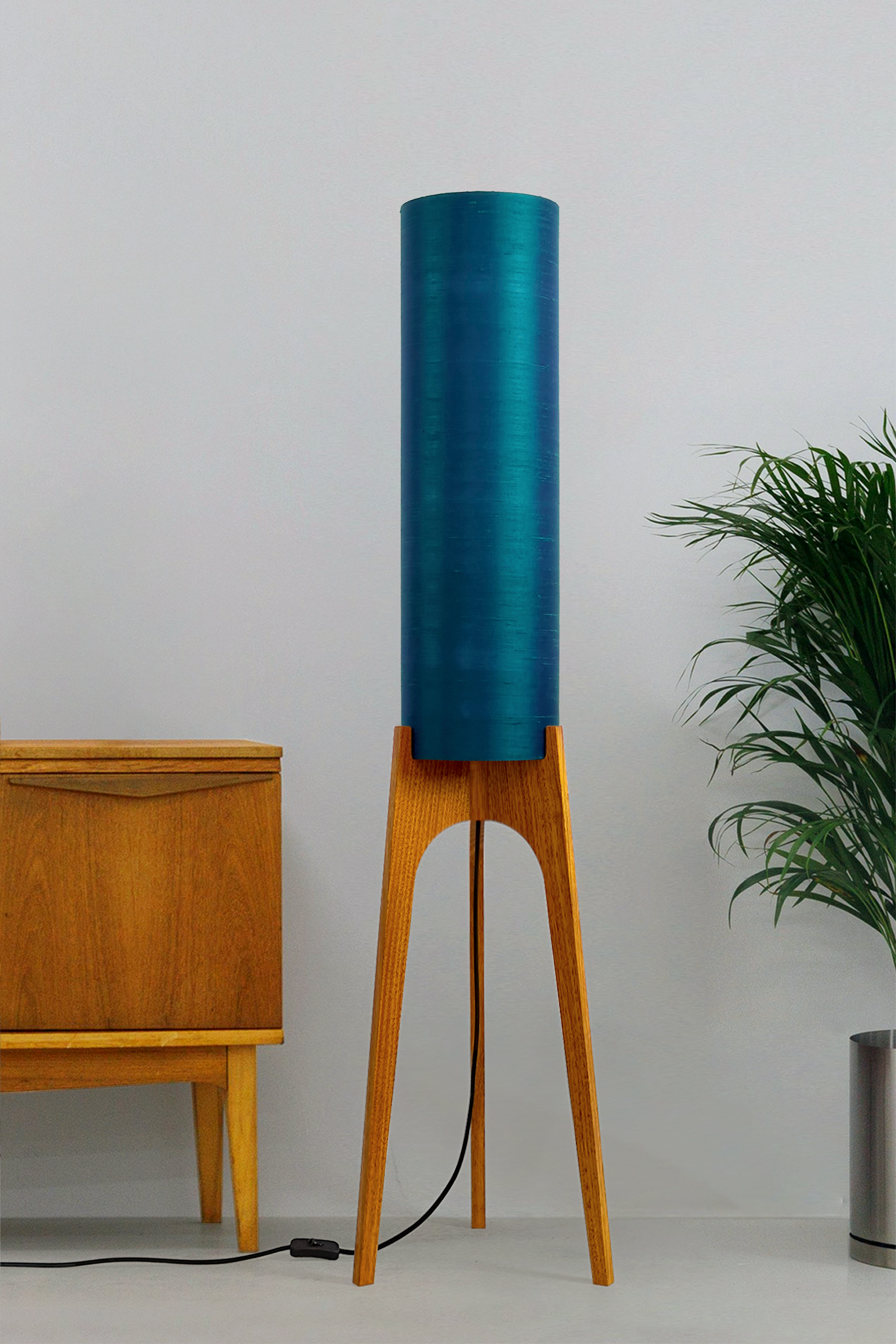 Wooden Rocket Floor Lamp with Teal Blue cylindrical handmade lampshade with premium raw silk, next to a wooden cabinet and plant on a light grey background