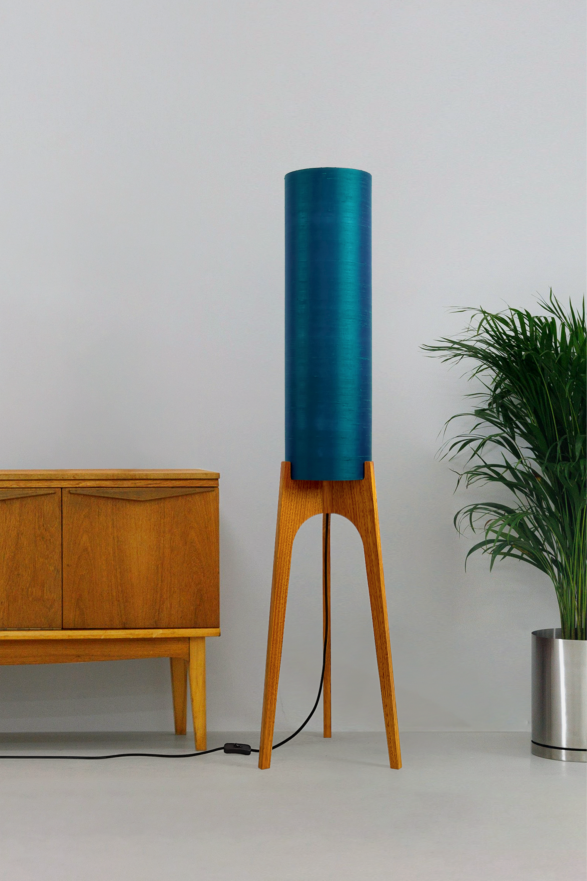 Wooden Rocket Floor Lamp with Teal Blue cylindrical handmade lampshade with premium raw silk, next to a wooden cabinet and plant on a light grey background