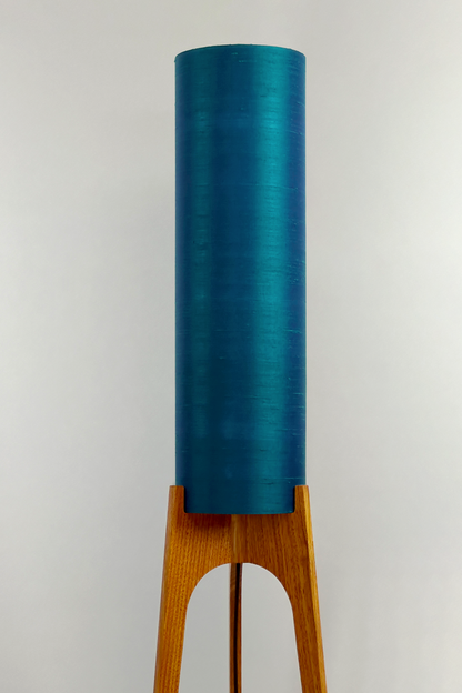 Wooden Rocket Floor Lamp with Teal Blue cylindrical handmade lampshade with premium raw silk, next to a wooden cabinet and plant on a light grey background