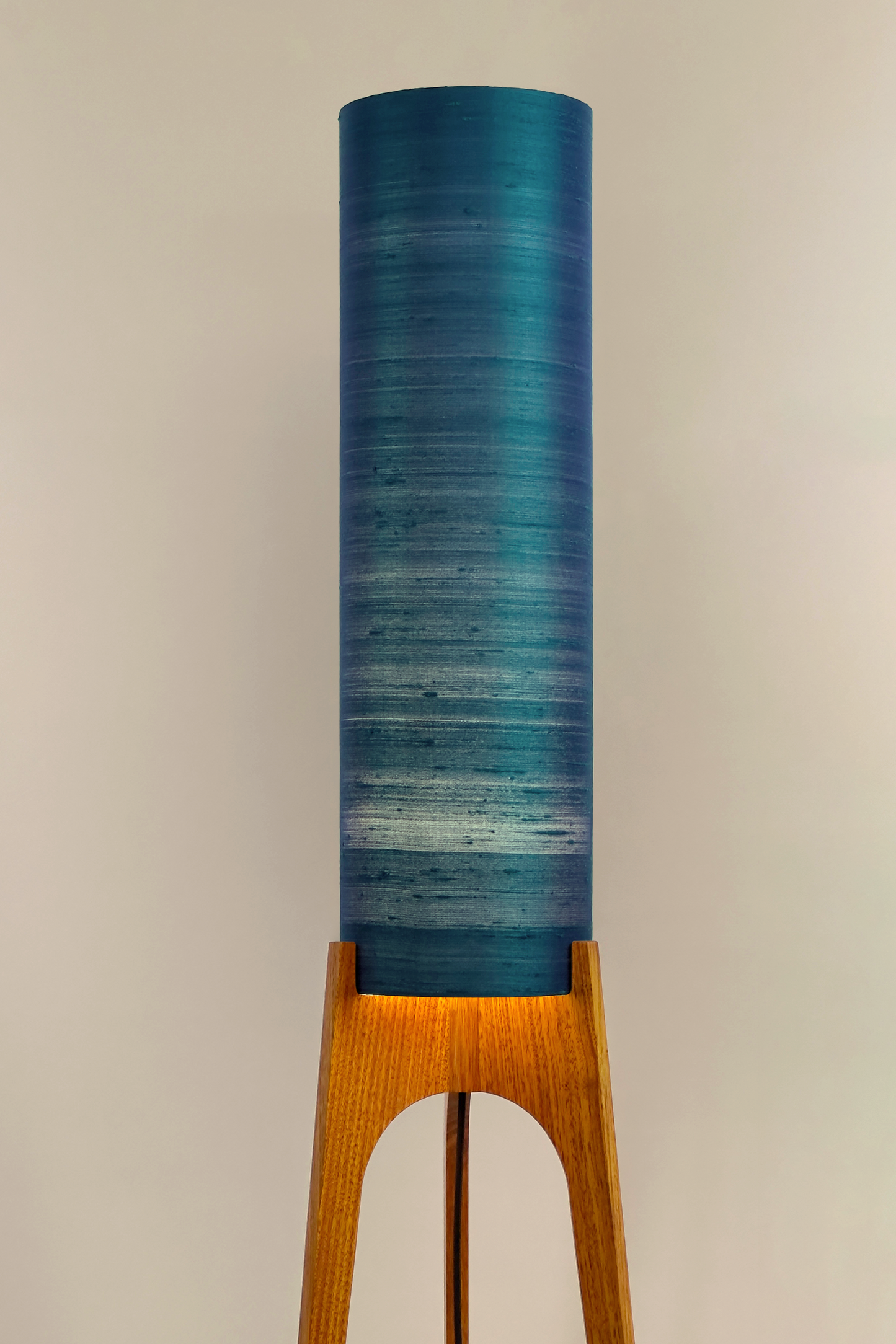 Wooden Rocket Floor Lamp with Teal Blue cylindrical handmade lampshade with premium raw silk, next to a wooden cabinet and plant on a light grey background