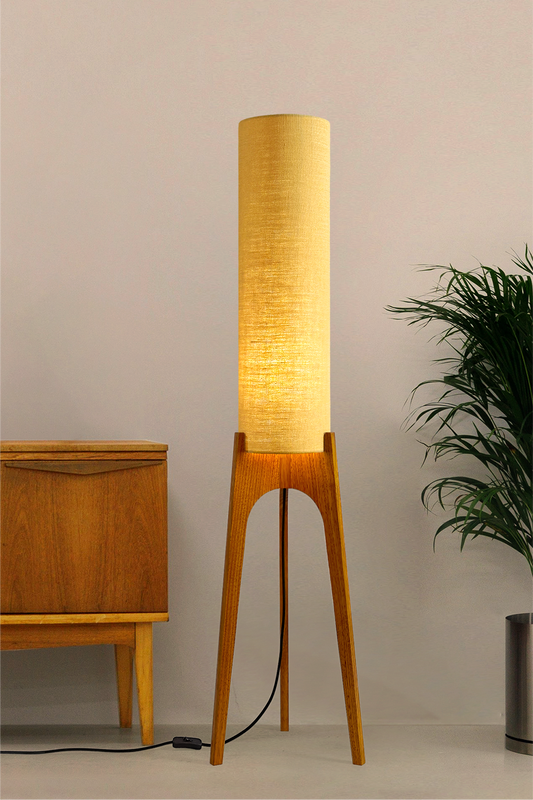 Wooden Rocket floor lamp with a premium linen shade in Wheat next to a wooden cabinet and plant on a beige background